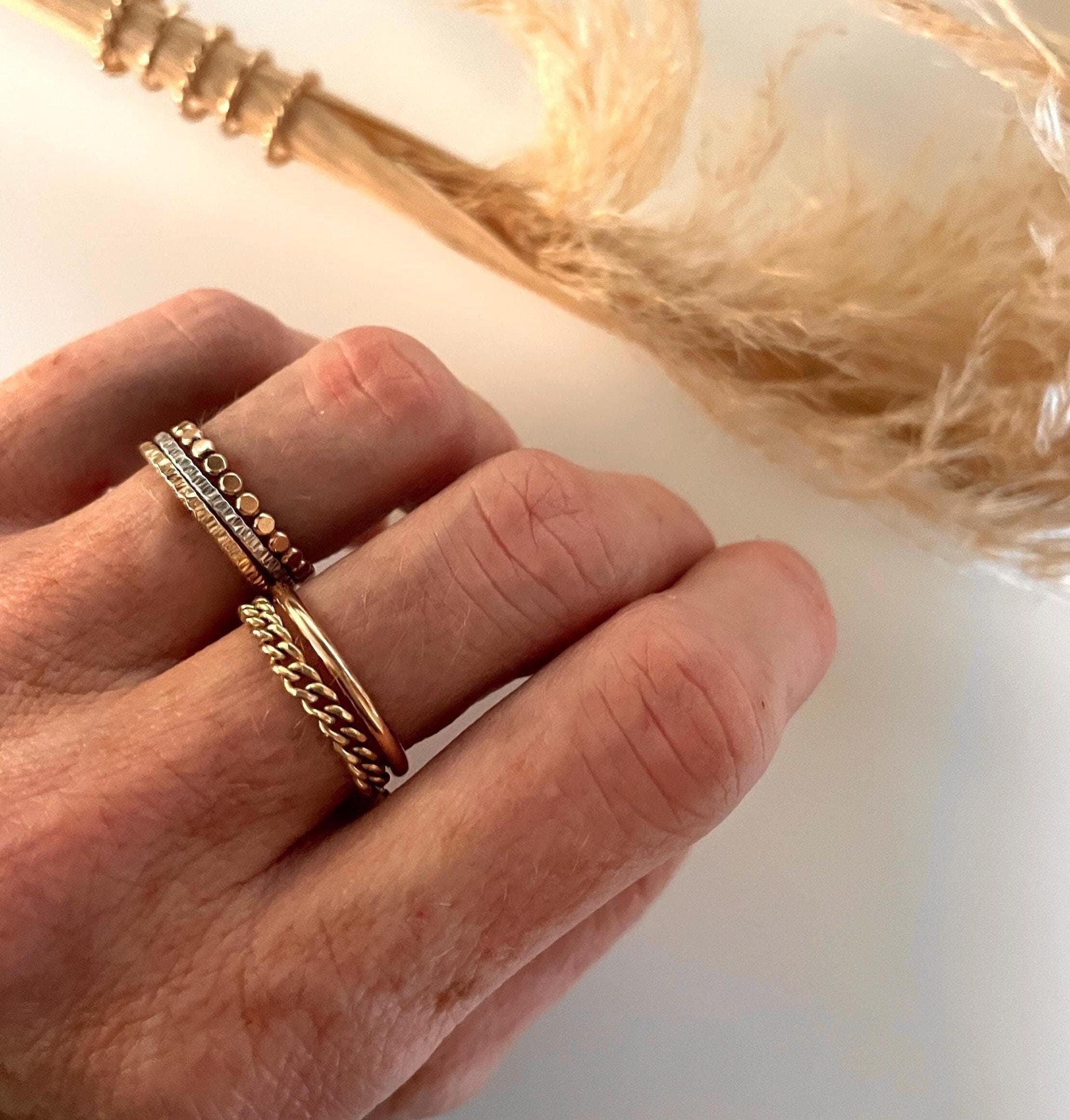 May include: Three gold rings stacked on a finger. The top ring is a thin band with small gold beads. The middle ring is a thin band with a hammered texture. The bottom ring is a thicker band with a twisted rope design.