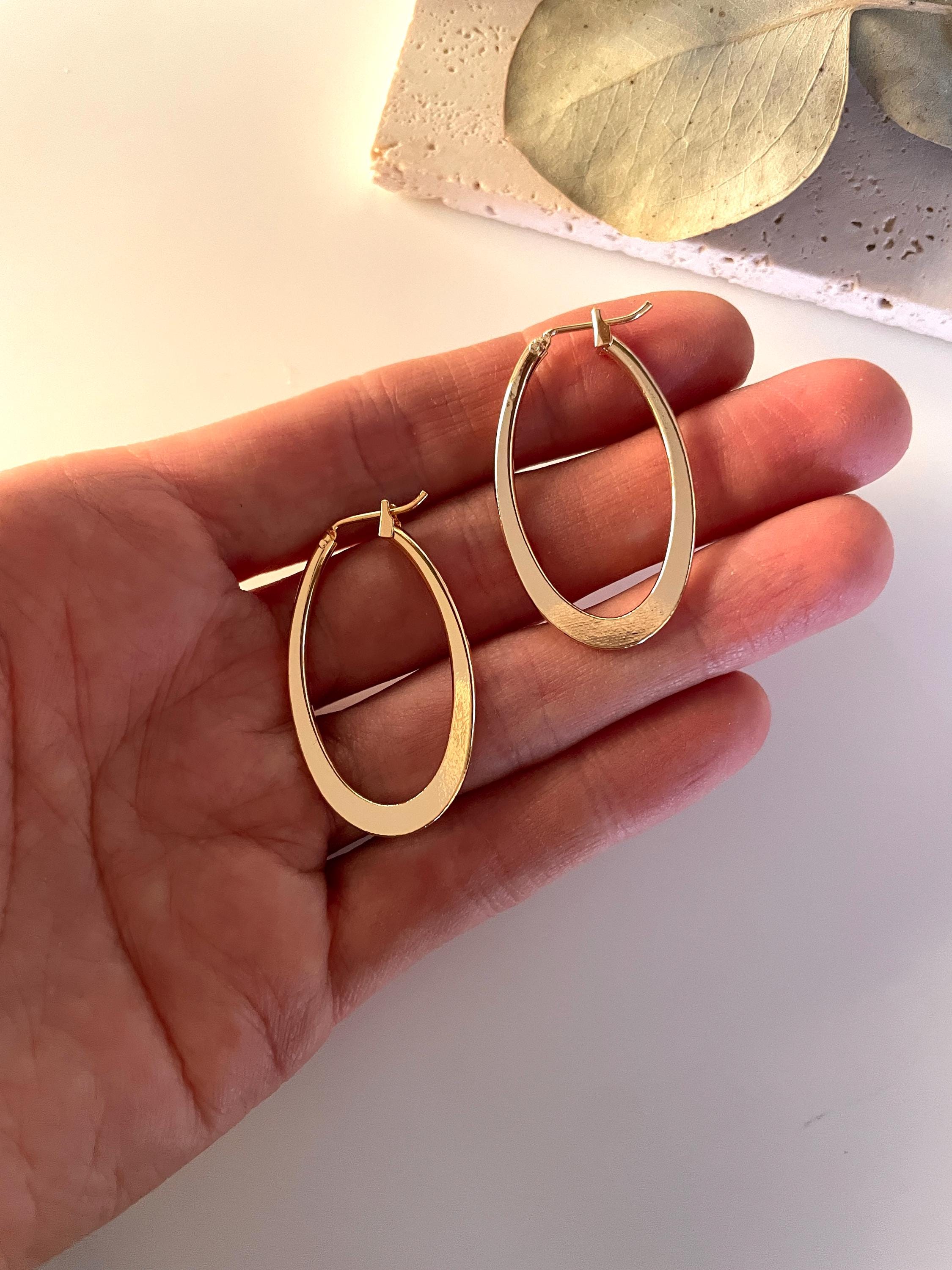 May include: A pair of gold-colored oval hoop earrings. The earrings are smooth and shiny, with a simple, minimalist design. They have a classic hoop shape with a post closure. The earrings are displayed against a neutral background, highlighting their metallic finish.